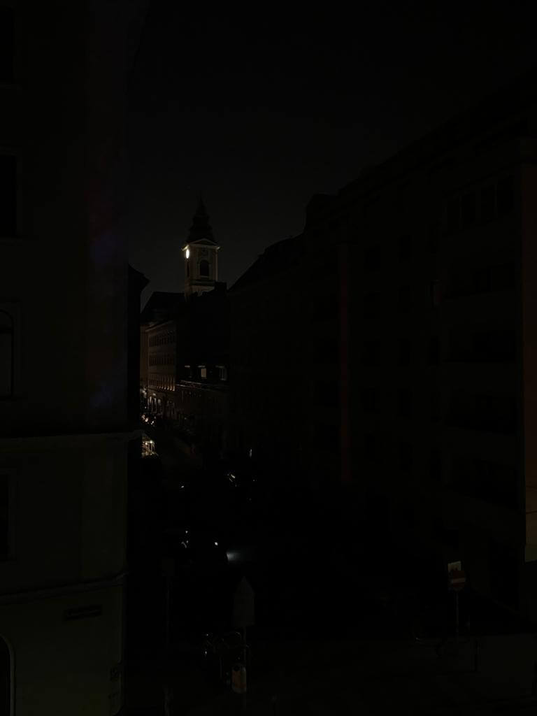 Blick aus dem Fenster einer Stadtwohnung im 3. Stock. Alles ist dunkel. Nur ein Kirchturm wird schwach beleuchtet und einzelne Lichter sind auf der Straße zu erkennen.